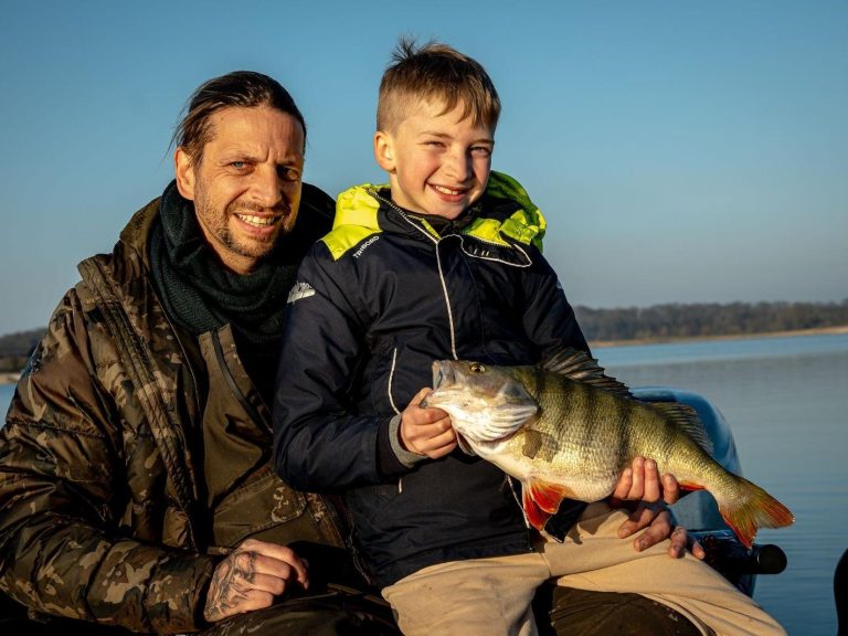 Vader en zoon tonen trots hun gevangen baars aan het water.