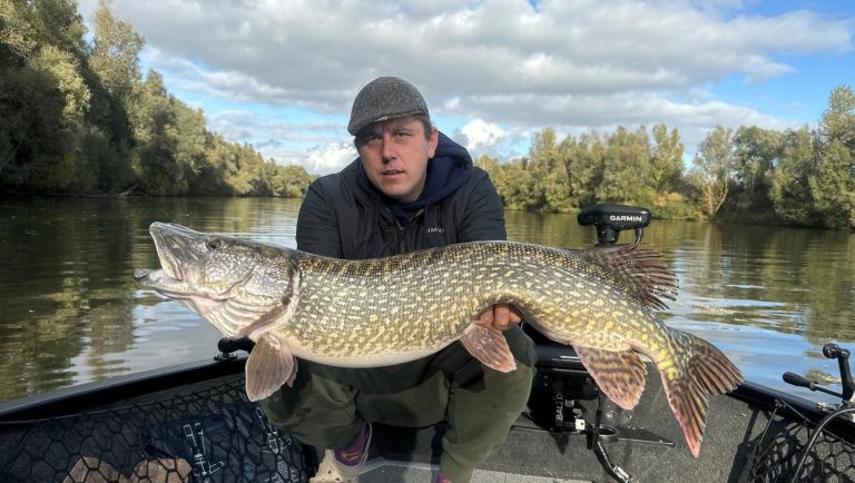 Man houdt grote snoek vast op een boot, met een waterachtige achtergrond.