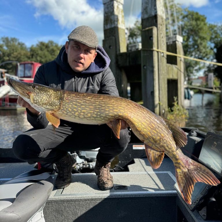 Man in een boot toont een grote snoek, met bomen en een brug op de achtergrond.