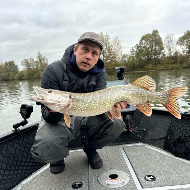 Een man op een boot houdt een grote snoek vast aan de oever van een meer.