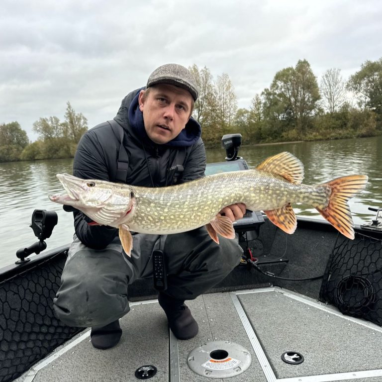 Man in een boot houdt een grote snoek vast, met een water- en bosachtige achtergrond.