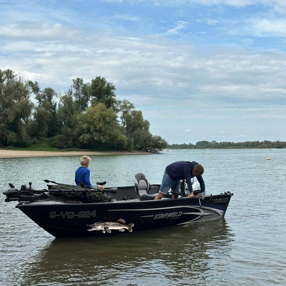 Visgids Dutch Fishing Experience Twee mensen aan het vissen in een boot op rustig water met bomen op de achtergrond.