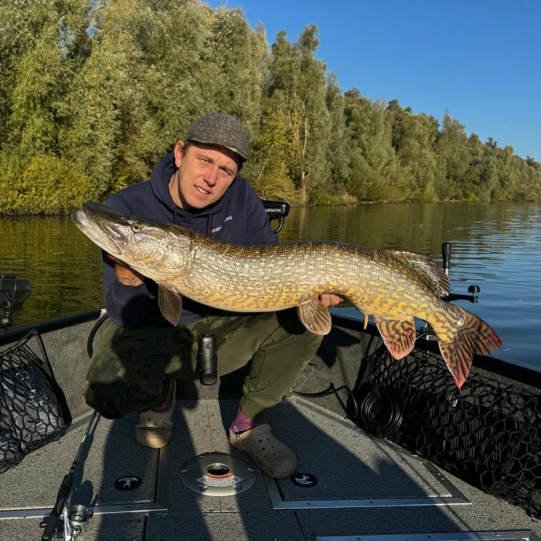 Man met een grote snoek op een boot in het water, met bewolkte lucht op de achtergrond.