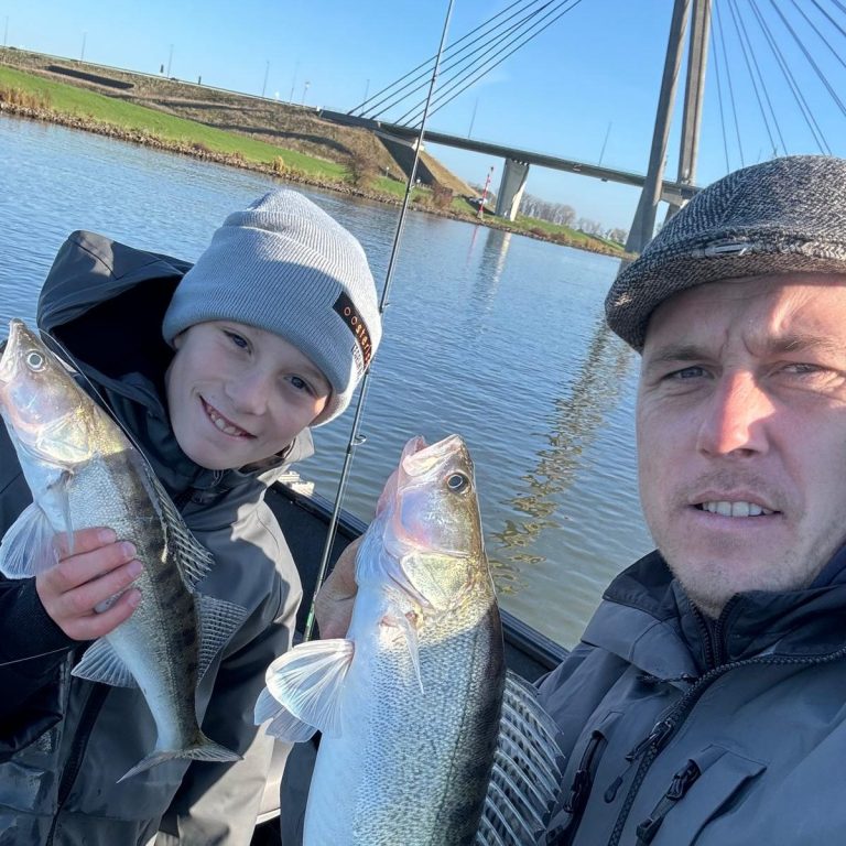 Kind en volwassene poseren met twee grote snoekbaarsen aan een waterkant met brug op de achtergrond.